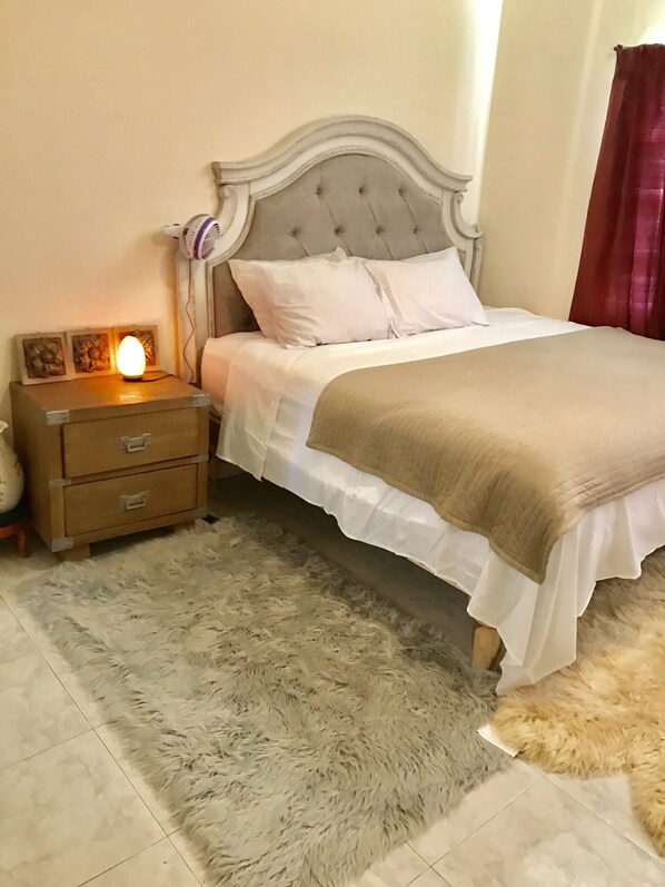 A neatly made bed with white bedding and a brown throw, a fluffy rug, and a bedside table with a lamp and decorative items.