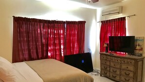 Een slaapkamer met een bed, een dressoir, een televisie en rode gordijnen.