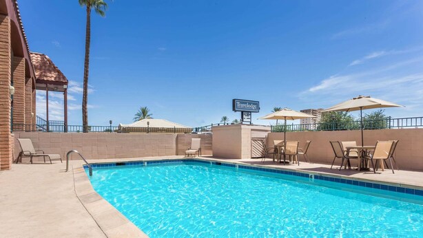 Une piscine avec des chaises longues et des parasols, une enseigne indiquant « Travelodge », et un ciel bleu azur.