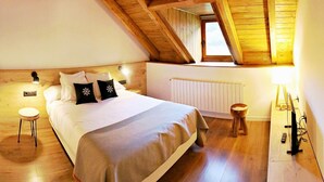 A cozy bedroom with a wooden ceiling, a large bed with white bedding and decorative pillows, a small bedside table, a nightstand, a radiator, and a wooden floor. - Lo Paller (Alt Aneu)