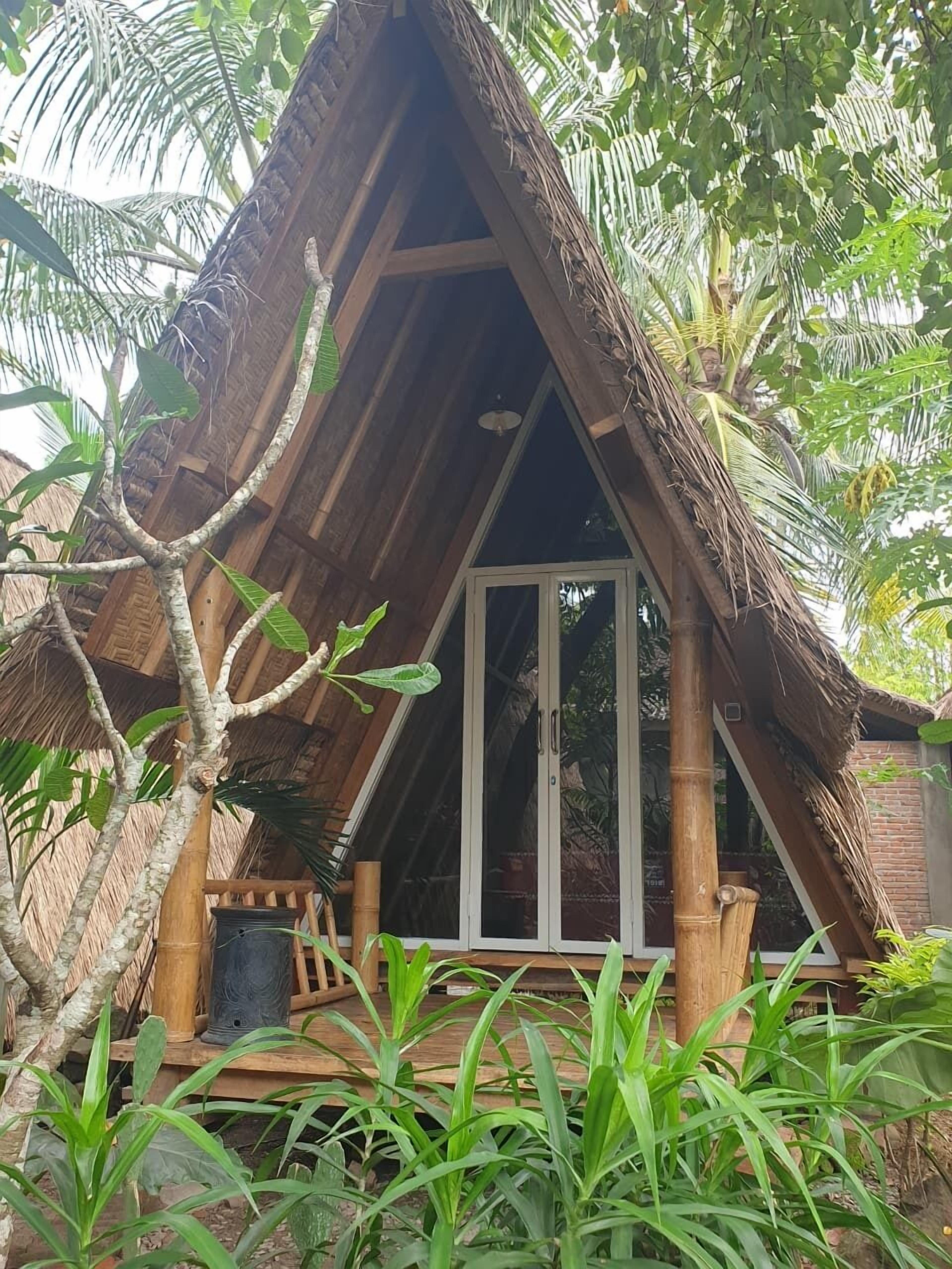 A thatched-roof hut with a glass door, surrounded by lush greenery.