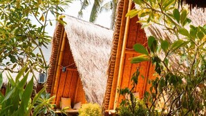 Una estructura con techo de paja, un banco de madera y una pequeña mesa, rodeada de vegetación tropical y un sendero de grava.