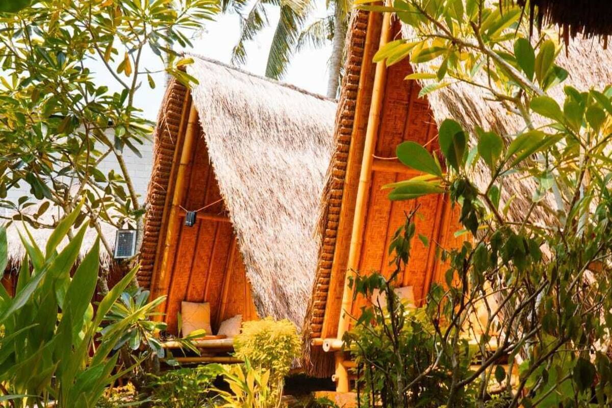 Una estructura con techo de paja, un banco de madera y una pequeña mesa, rodeada de vegetación tropical y un sendero de grava.