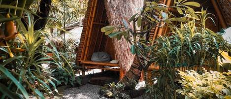Una cabaña con techo de paja y un área de estar con cojines, rodeada de densa vegetación tropical y un sendero de grava.