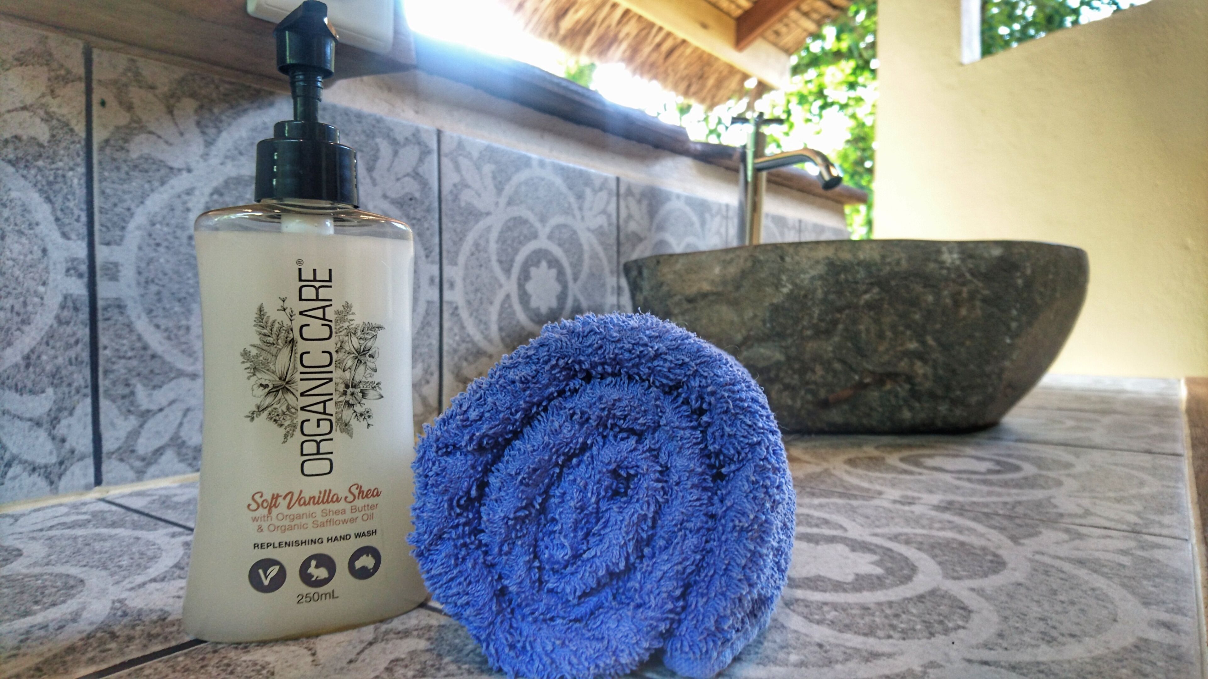 Un flacon de beurre de karité à la vanille douce de la marque Organic Care, enrichi en huile de carthame biologique et en savon pour les mains biologique, une serviette bleue et un lavabo en pierre.