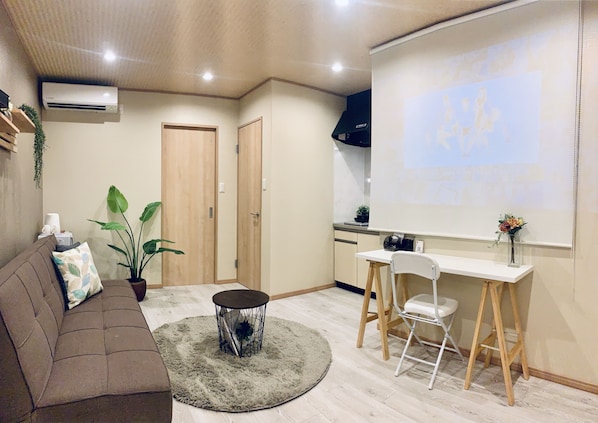 A modern living room with a brown sofa, a coffee table, and a dining area with a white table and chairs. There is a projector screen on the wall and a kitchen area in the background.