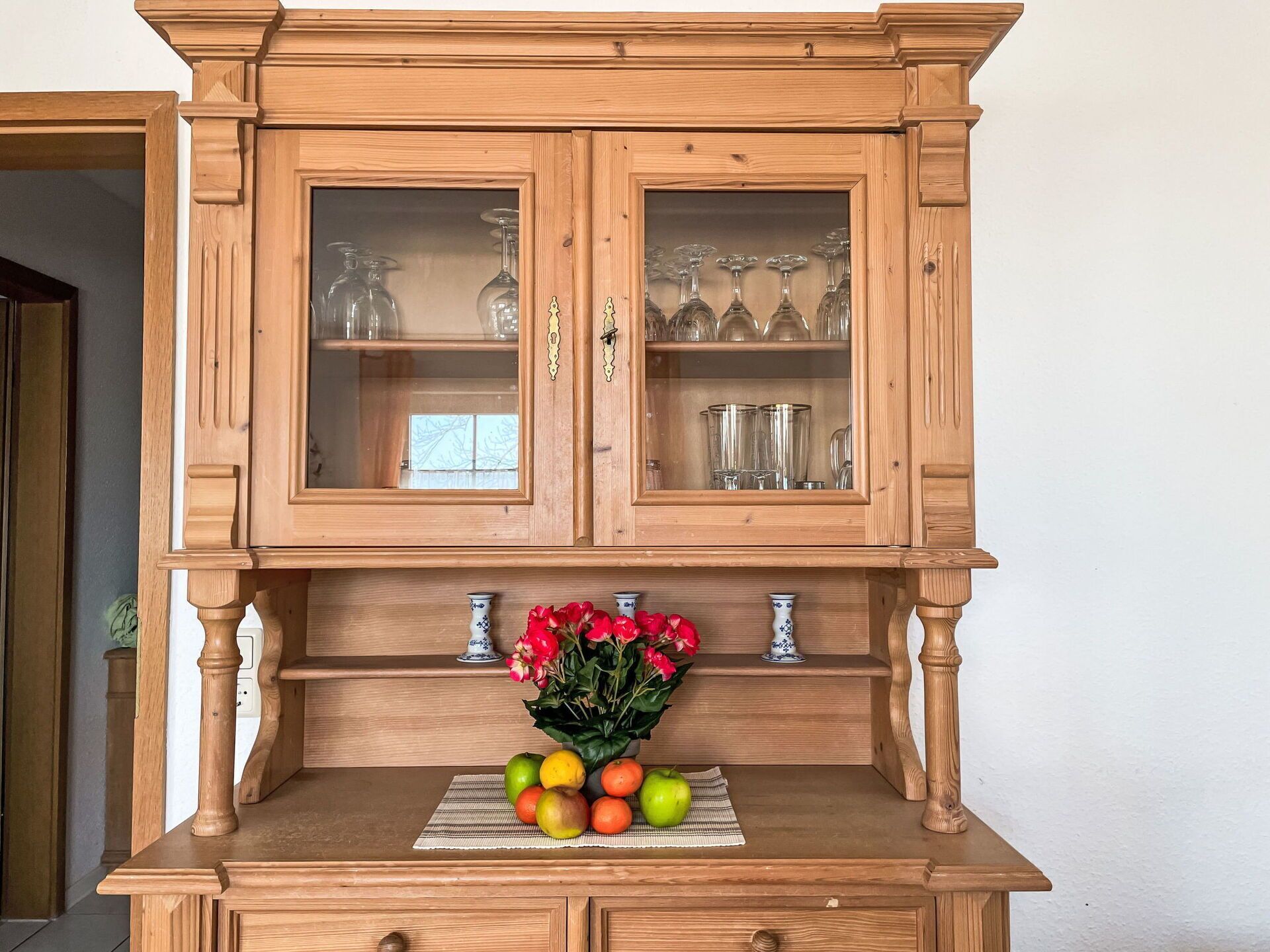 A wooden cabinet with glass doors, a fruit bowl with apples and oranges, and a vase with red flowers.