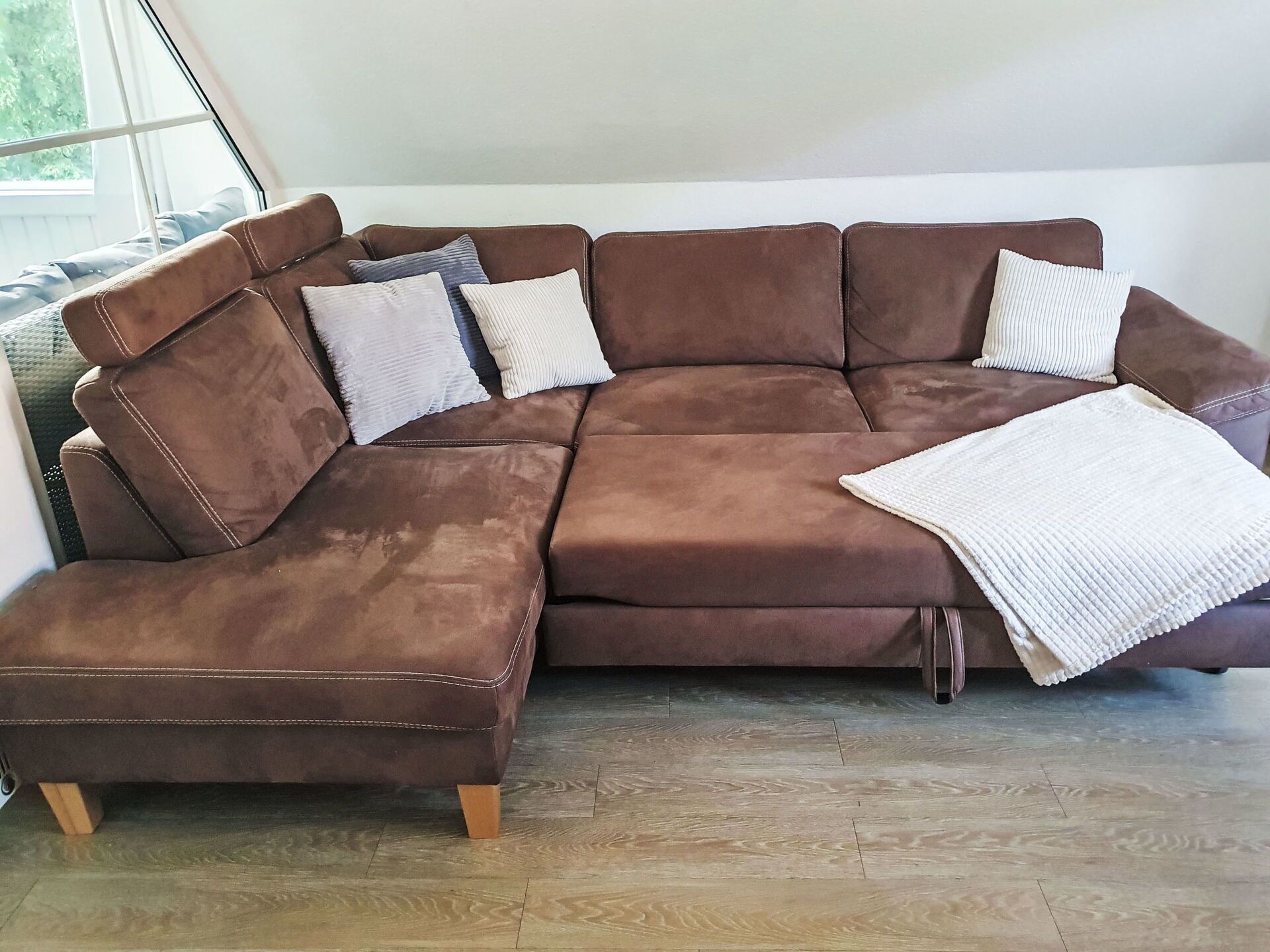 A brown sectional sofa with a white throw blanket and multiple pillows in a room with wooden flooring.