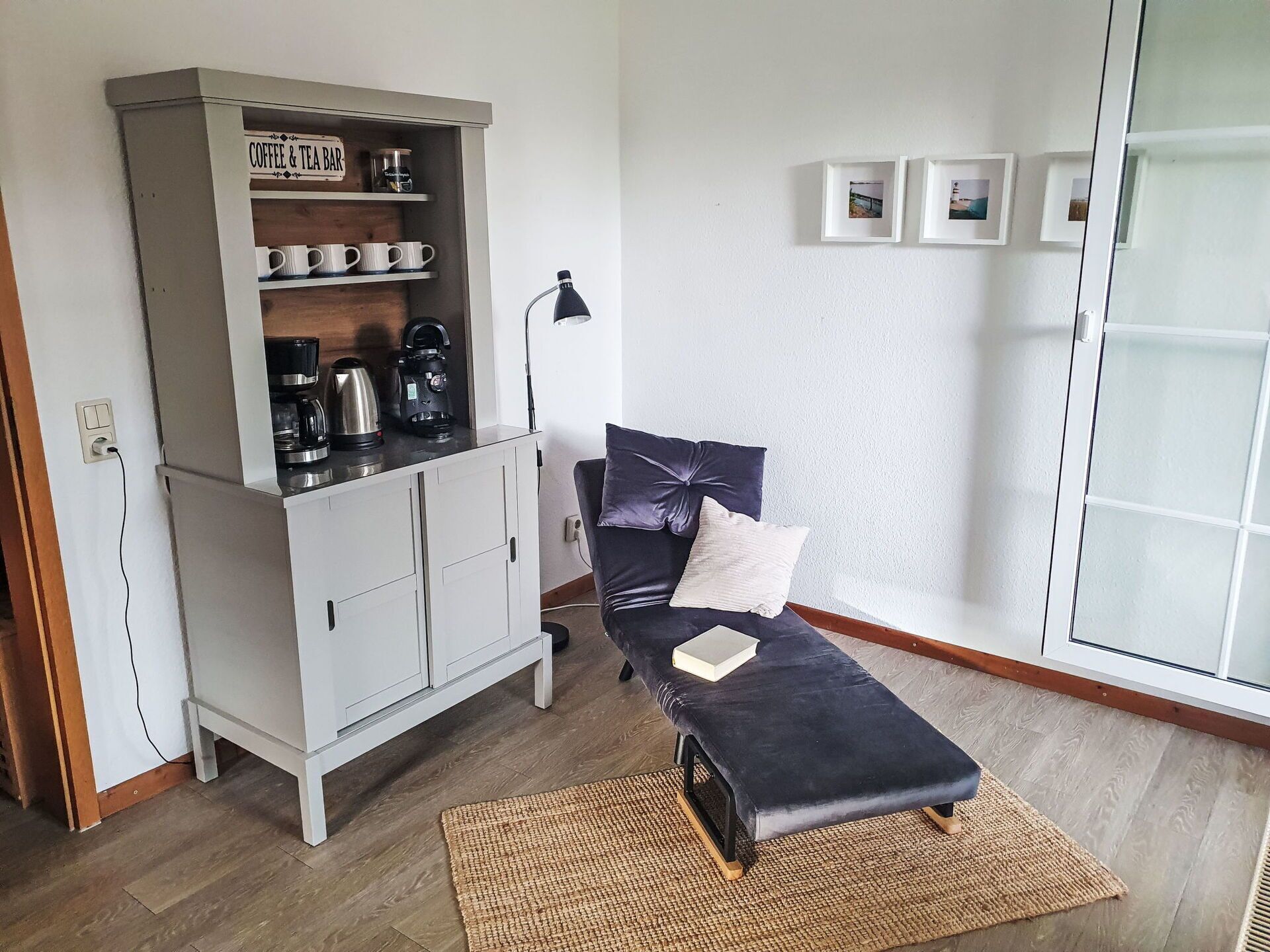 A compact room with a bed, a cabinet with coffee and tea items, a lamp, and a wall with framed pictures.