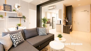 A modern living room with a grey sectional sofa, a round coffee table, and decorative pillows. There is a kitchen area visible in the background with cabinets and a countertop. - The Robertson Suites Bukit Bintang (Kuala Lumpur)