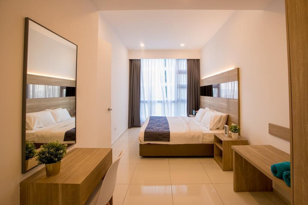 A modern hotel room with a large bed, a bedside table, a mirror, and a view of the outdoors through large windows.