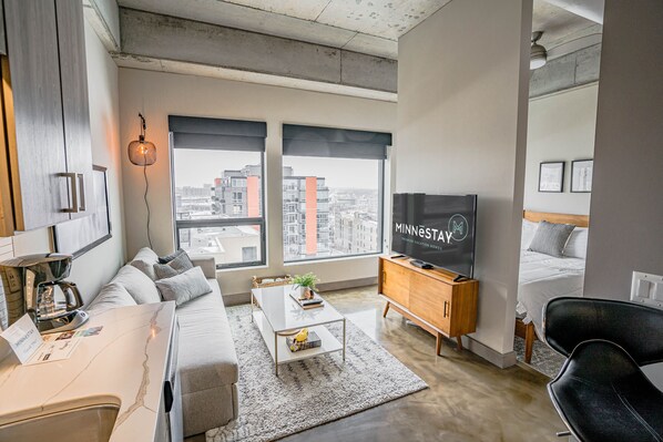 A modern hotel room with a large bed, a TV, a sofa, a coffee table, and a view of the city through large windows.