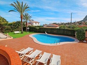 A swimming pool with a curved edge, surrounded by a tiled patio. There are white lounge chairs and a white bench.