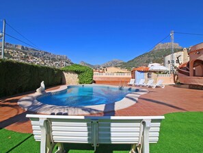 A swimming pool with a white bench in the foreground, surrounded by a patio area with lounge chairs and a small building with a tiled roof.