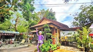 Una persona vestida de color morado parada frente a un edificio con techo de tejas y base de ladrillo, rodeada de plantas en macetas y mobiliario de exterior.