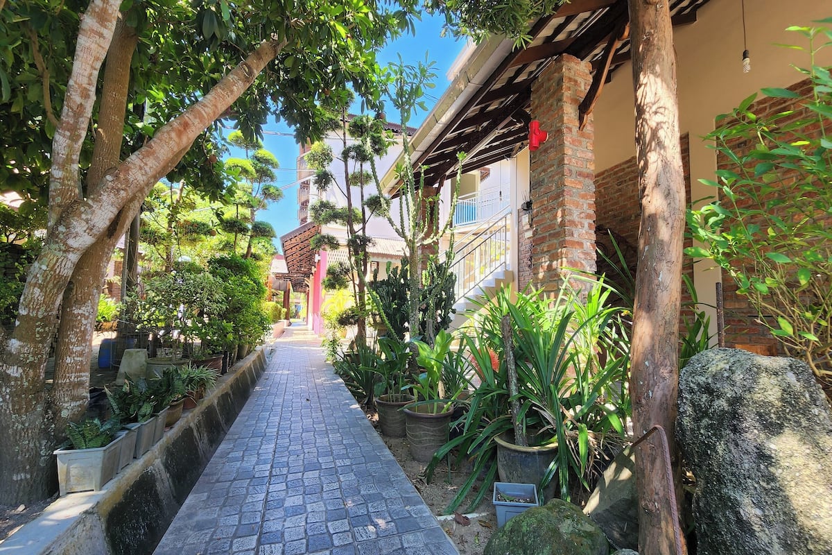 A narrow pathway flanked by potted plants and trees, leading to a building with a brick facade and a balcony.