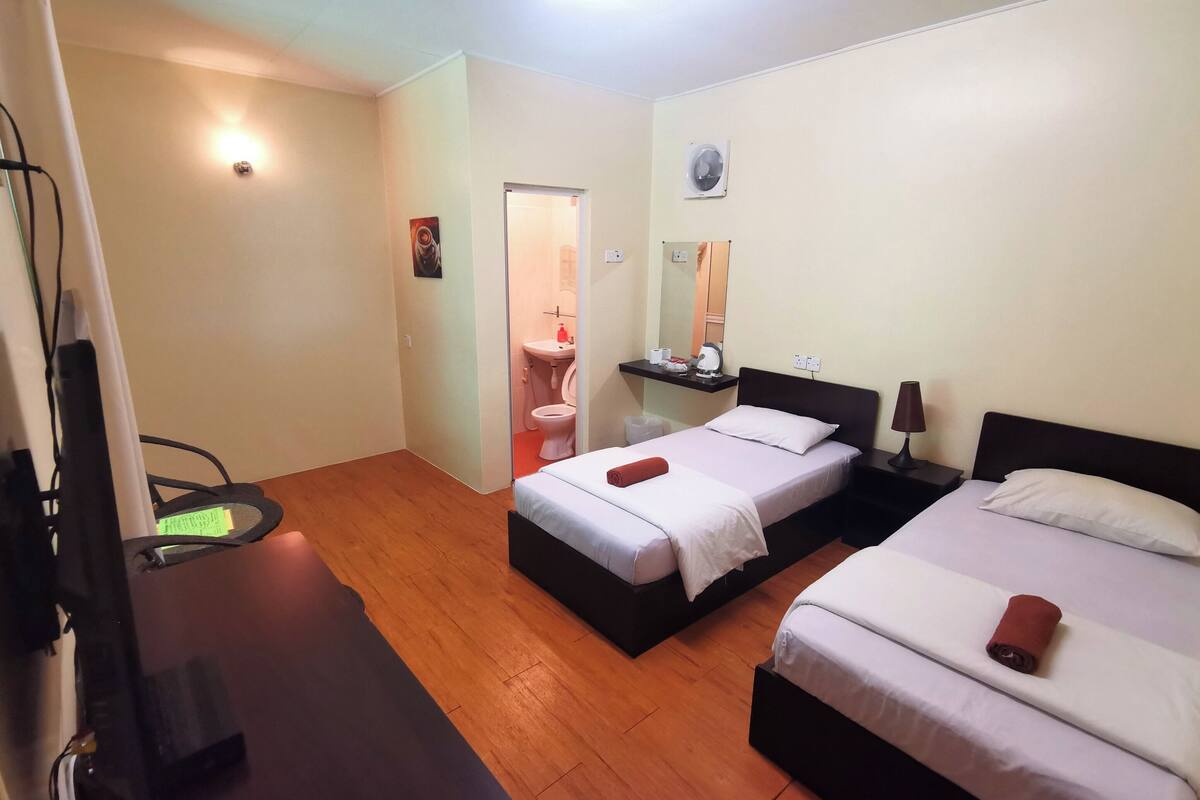 A hotel room with two beds, a desk, a chair, a bathroom, and a small shelf with toiletries.