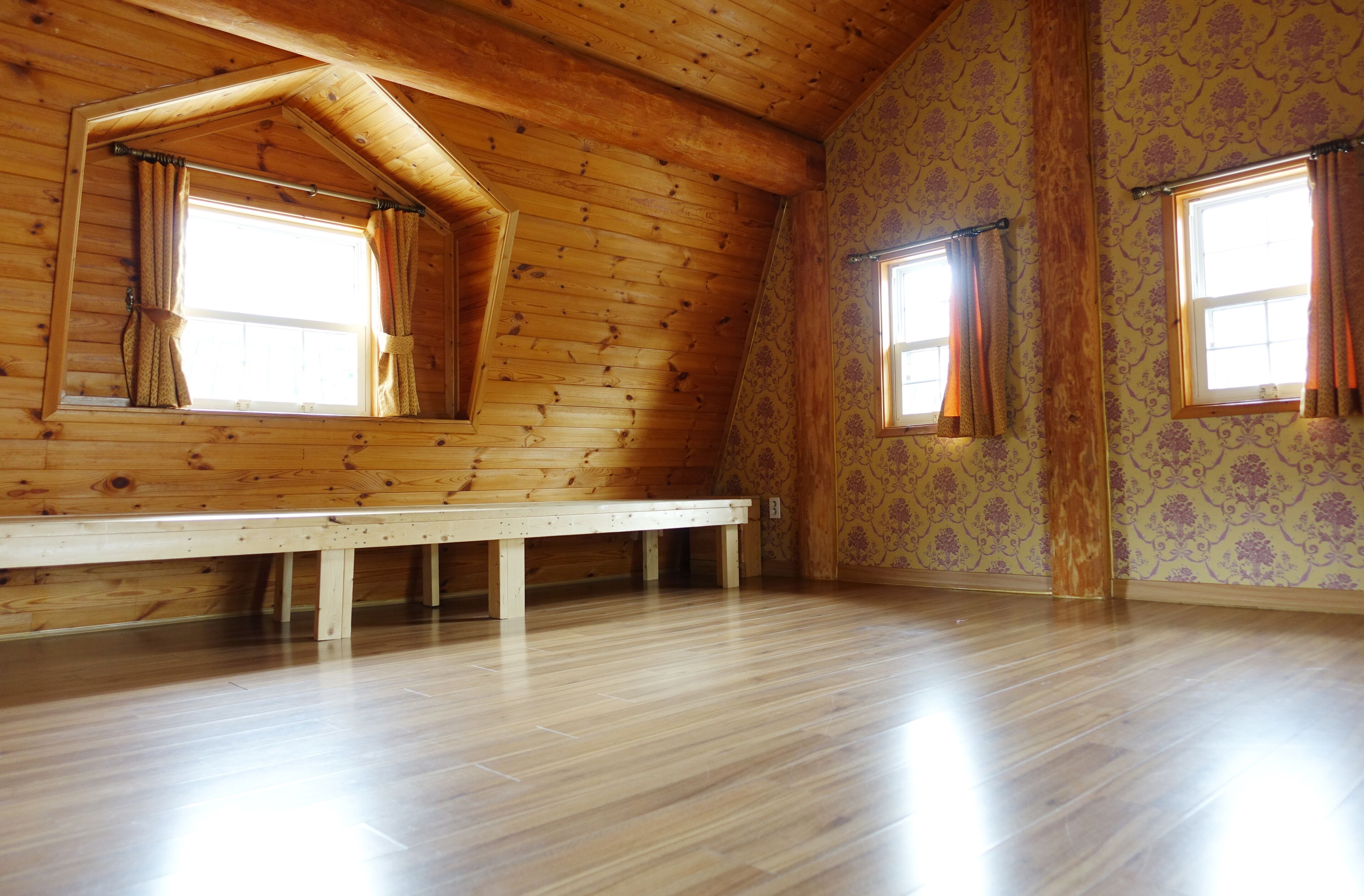 A room with wooden walls, a sloped ceiling, and a bench. There are two windows with curtains.