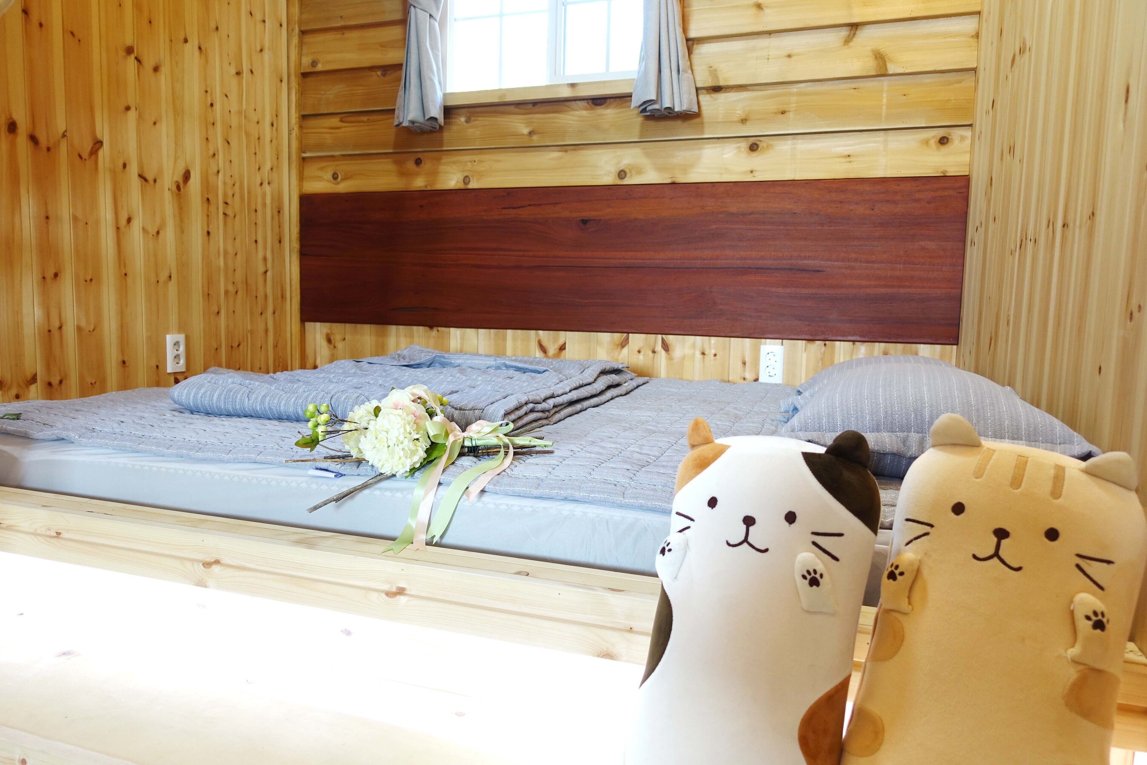 Una habitación tipo cabaña de madera con una cama, un ventanal con cortinas y dos almohadas con forma de gato.