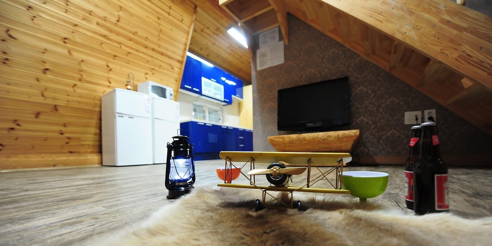 Ett mysigt rum med träväggar, ett litet pentry, en tv, ett leksaksflygplan, en lykta och två ölflaskor.