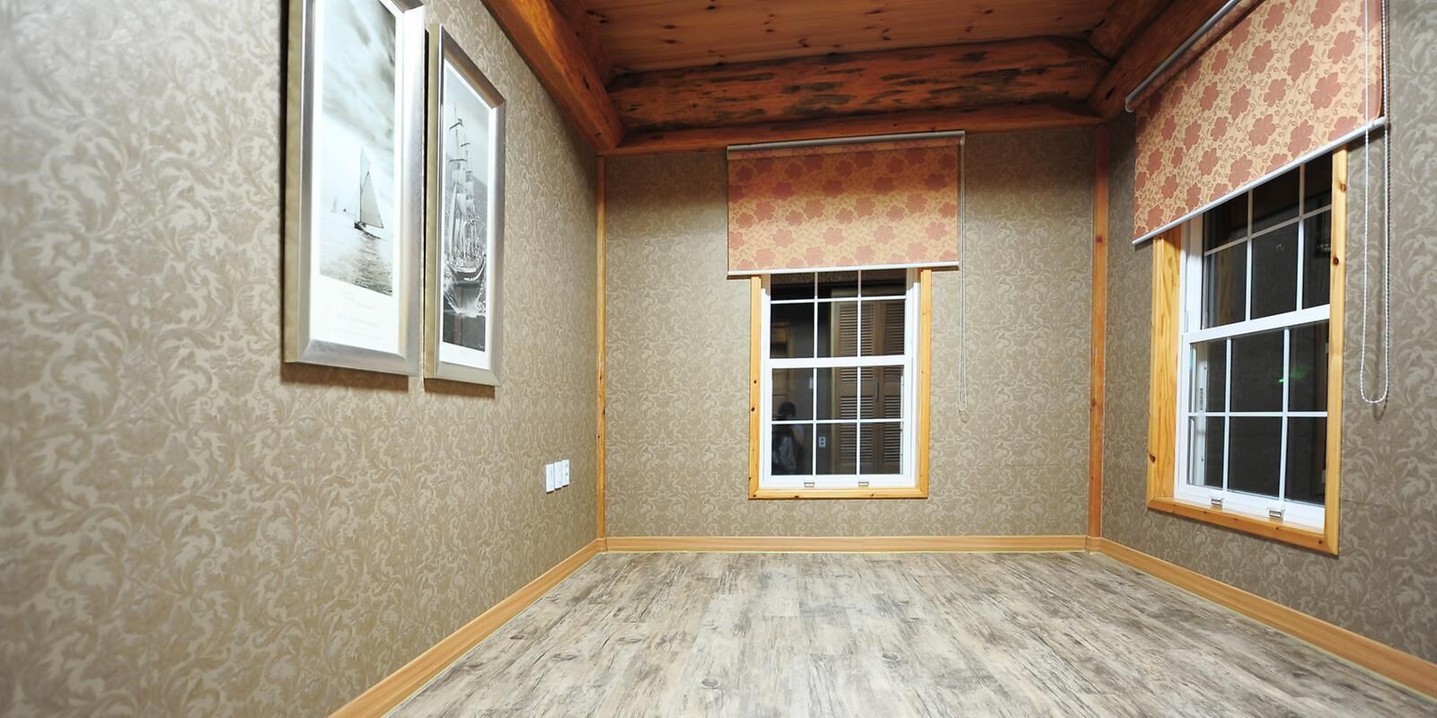 A room with patterned wallpaper, wooden flooring, and two windows with blinds.