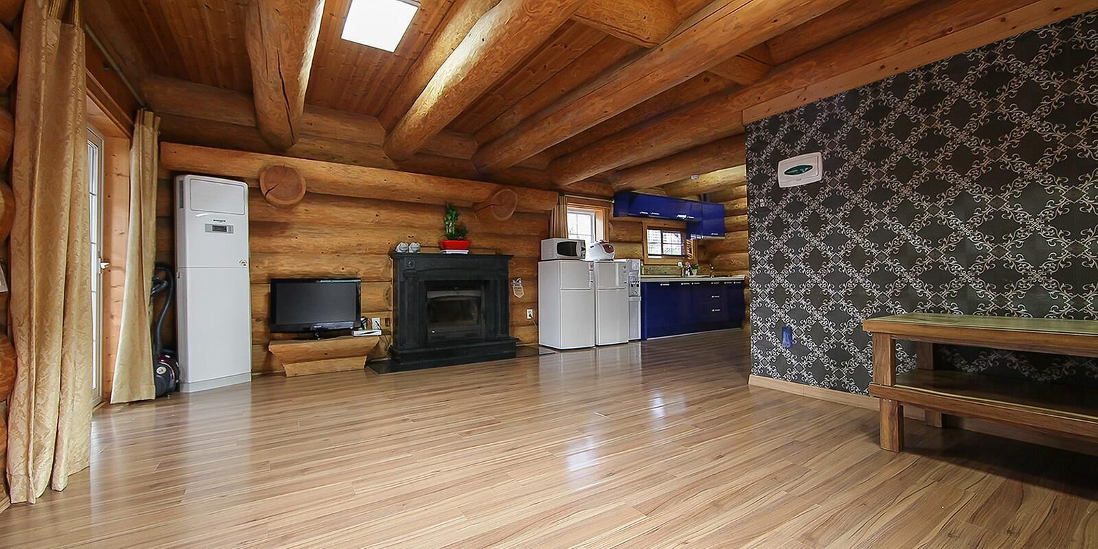 Interior de una cabaña de troncos con piso de madera, chimenea, televisor, zona de cocina y pared con diseño.