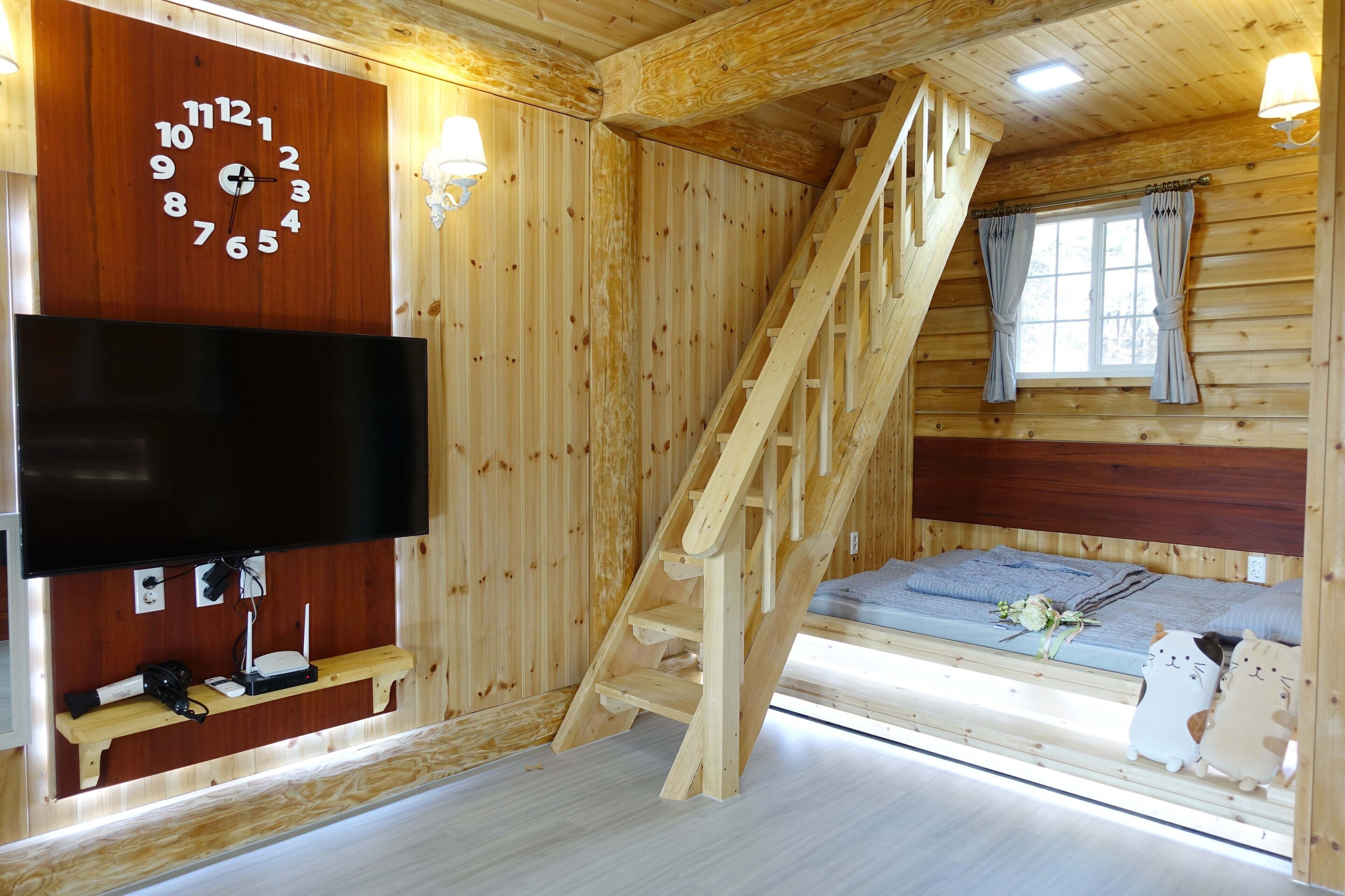 A wooden cabin interior with a wooden staircase, a bed with a window, a wall-mounted TV, and a shelf with electronic devices.