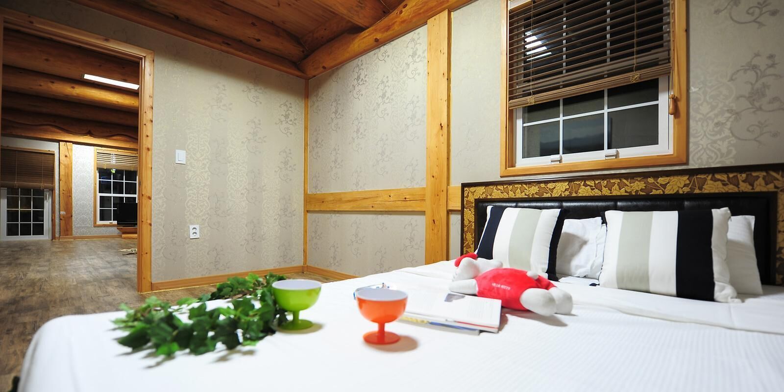 A hotel room with a bed, a window with blinds, a bedside table with a book and a glass, and a mirror on the wall.