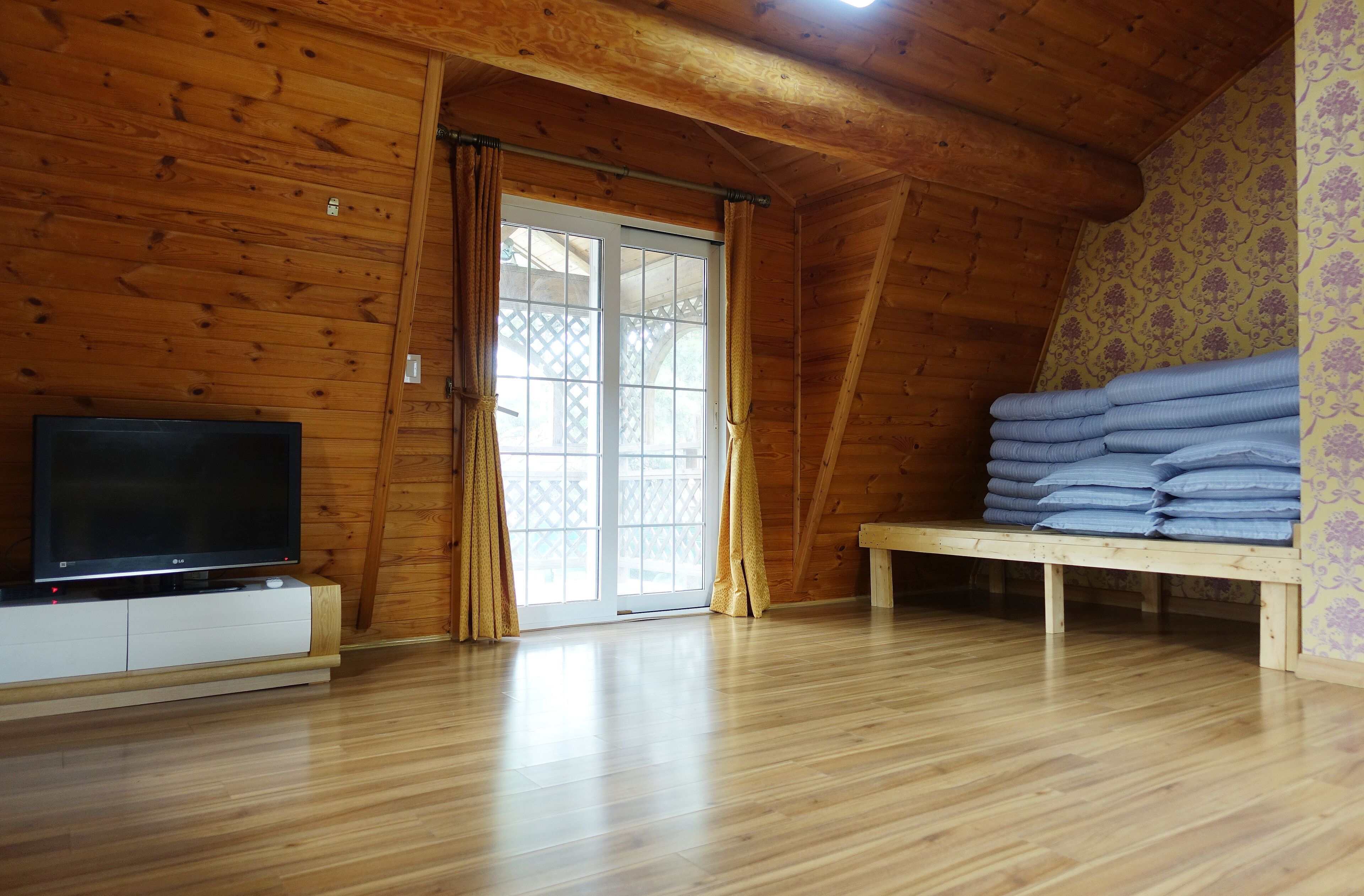 A wooden-paneled room with a TV on a white cabinet, a bench with folded blankets, and a sliding glass door to the outside.