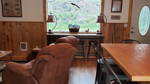 A cozy room with a wooden ceiling, a large window with a view of greenery, a brown leather chair, a wooden table, and a lamp. - Hunting ~n~ Hooks Getaway (Salmon)
