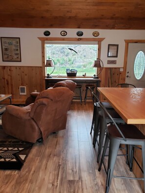 A cozy room with a wooden ceiling, a large window with a view of greenery, a brown leather chair, a wooden table, and a lamp.
