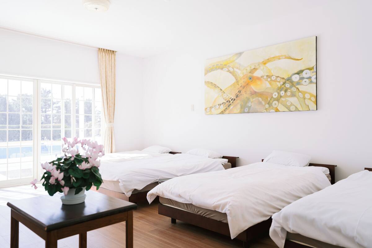A hotel room with three beds, a large window, and a painting on the wall.