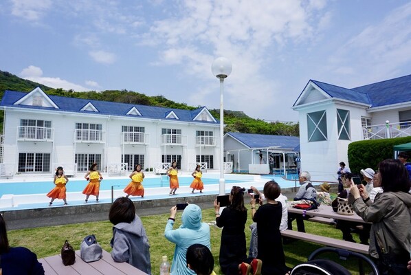 Un groupe de danseurs se produisant sur une scène devant un bâtiment doté d’une piscine.