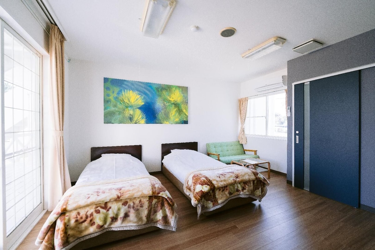 A hotel room with two beds, a large window, a green chair, and a painting on the wall.