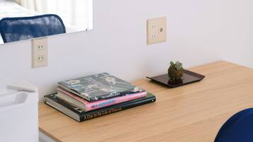 A hotel room with a desk, a flat-screen TV mounted on the wall, a mirror, and a small potted plant.