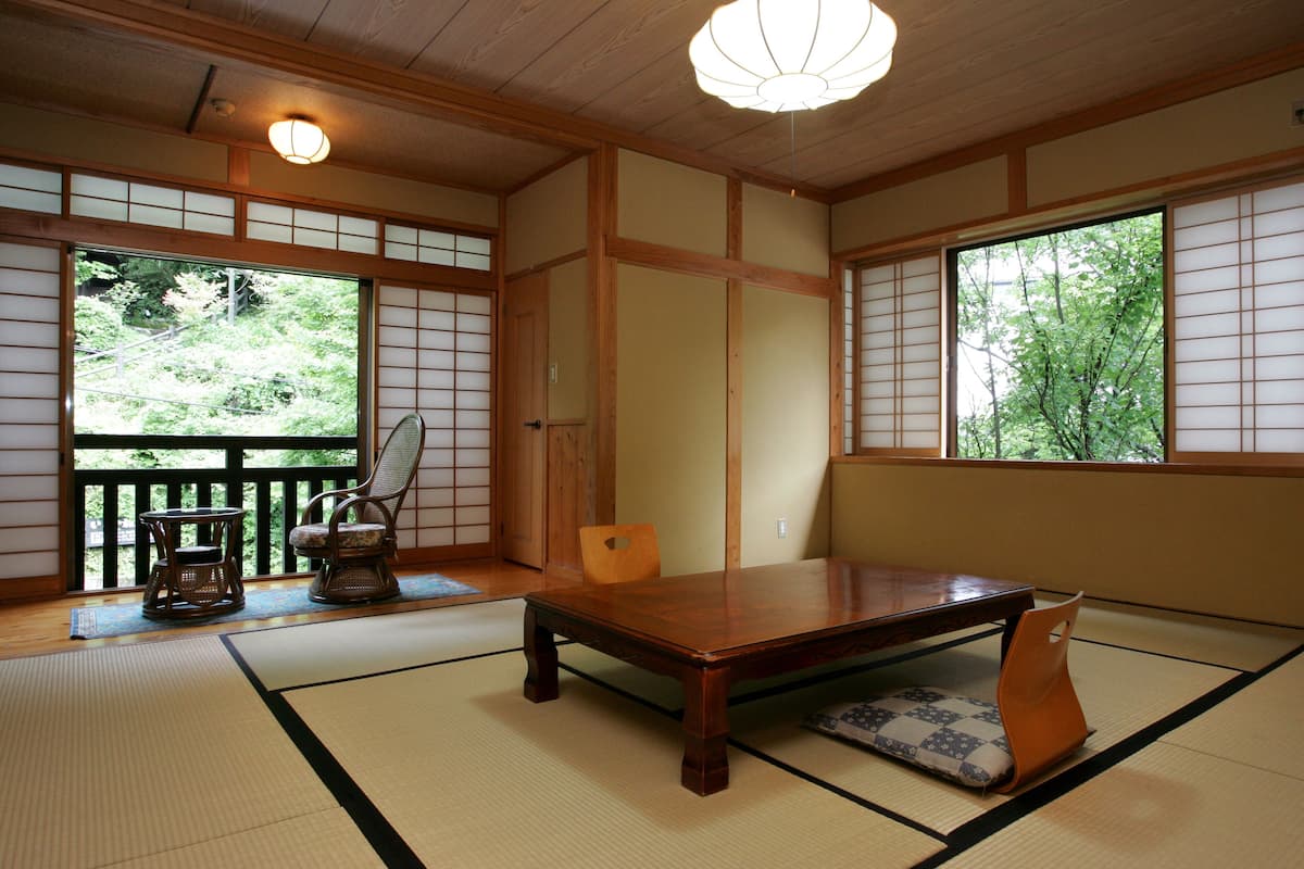 A traditional Japanese room with tatami flooring, a wooden table, a low table with a cushion, and a view of greenery through large windows.