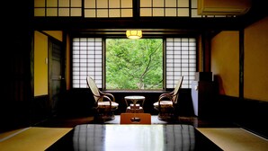 Una habitación japonesa tradicional con suelo de tatami, una mesa baja y dos sillas. Se ve un ventanal con vistas a la vegetación.