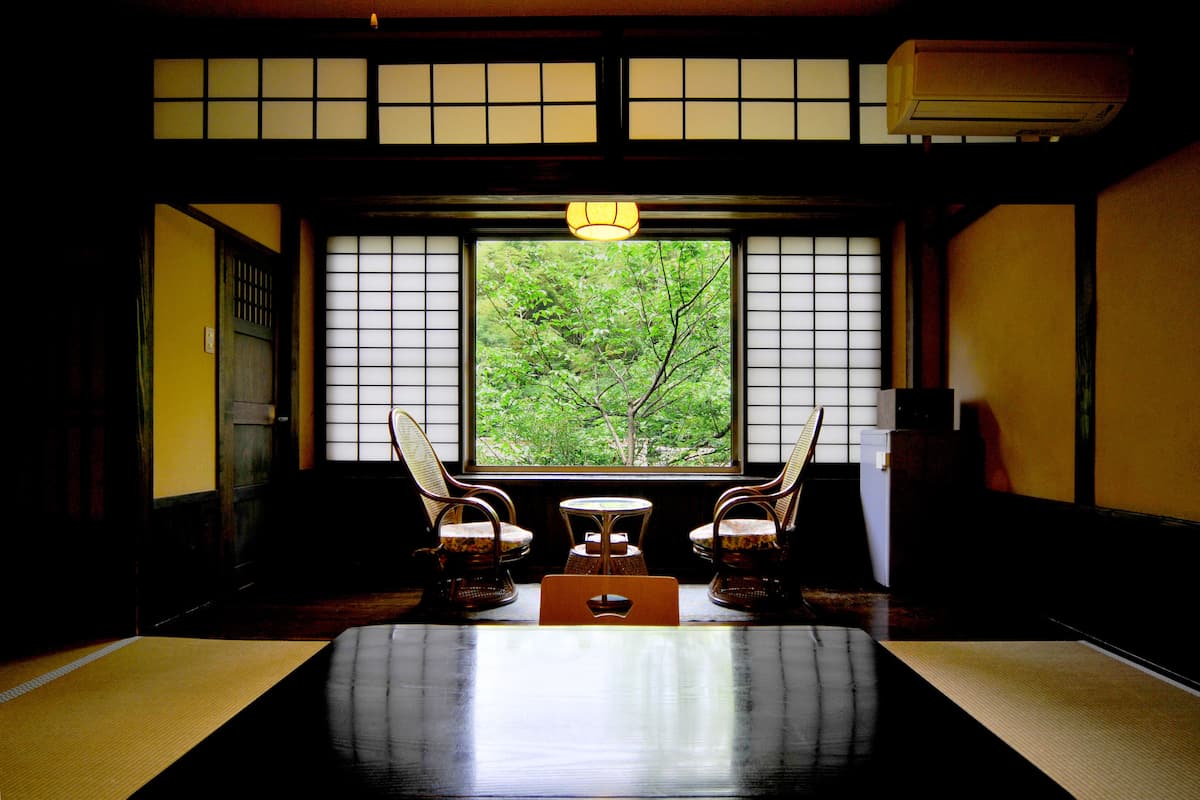 A traditional Japanese room with tatami flooring, a low table, and two chairs. A large window with a view of greenery is visible.