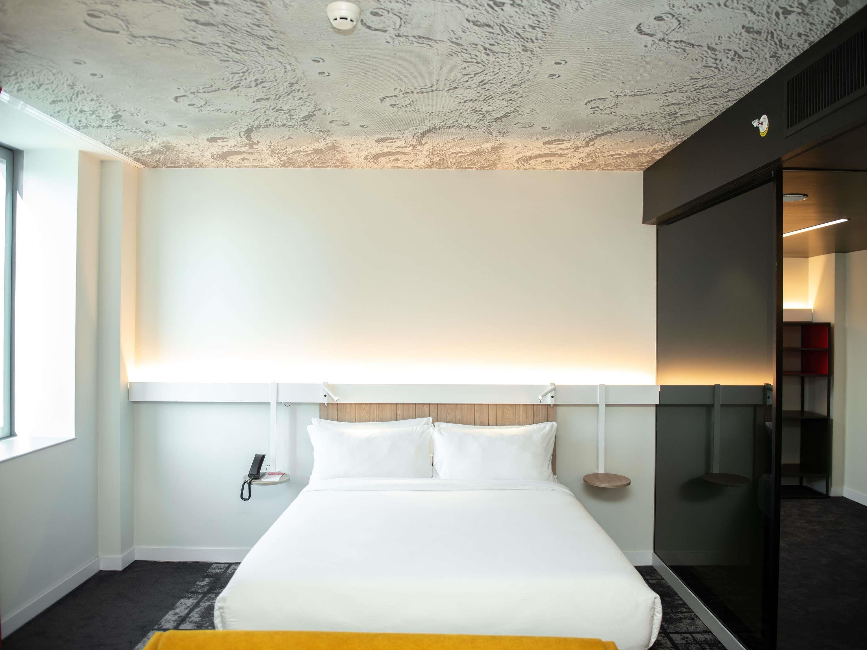 A modern hotel room with a large bed, a phone on the wall, and a glass partition.
