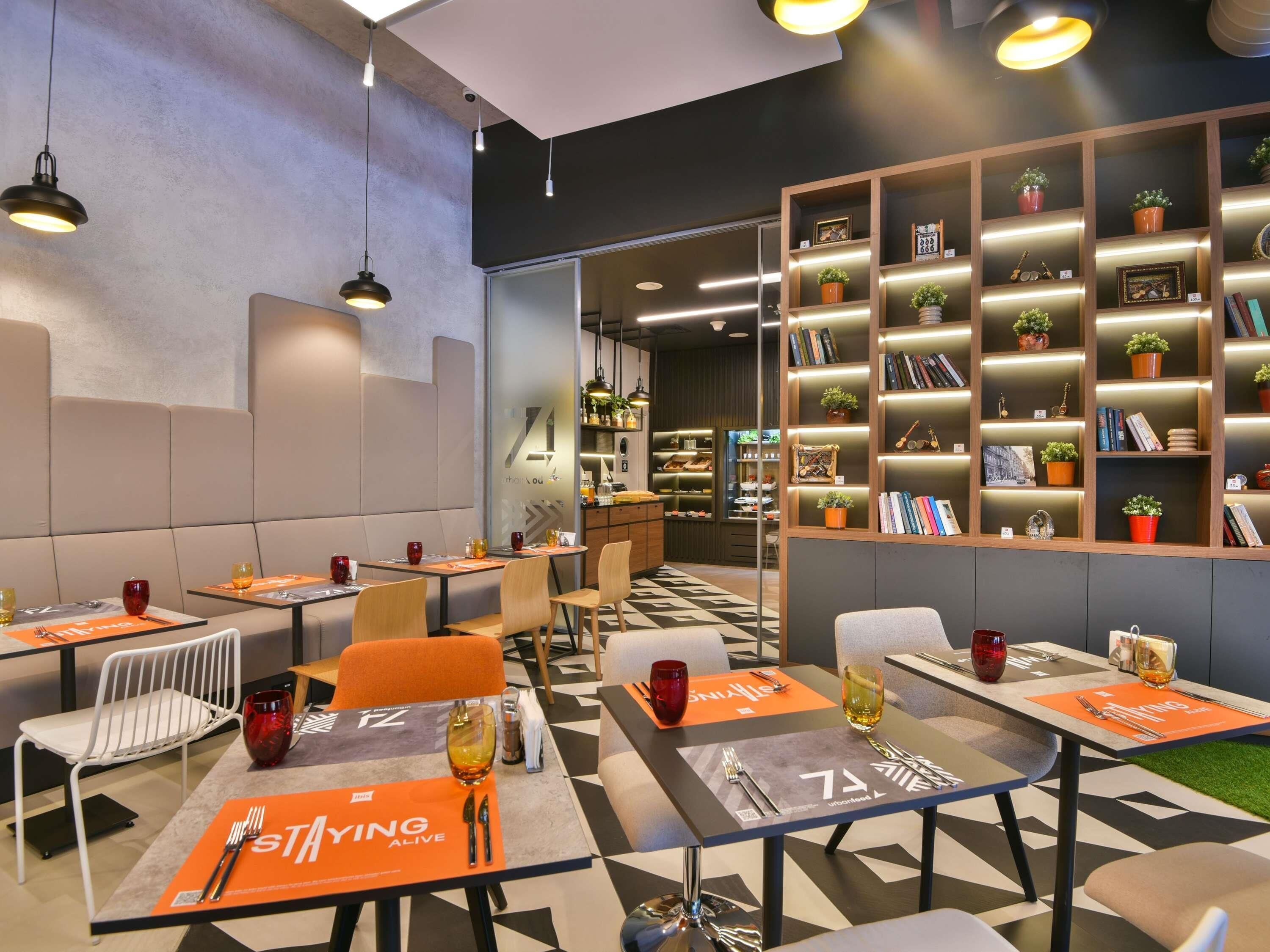 A modern restaurant with tables set for dining, a bookshelf with books and plants, and a bar area.