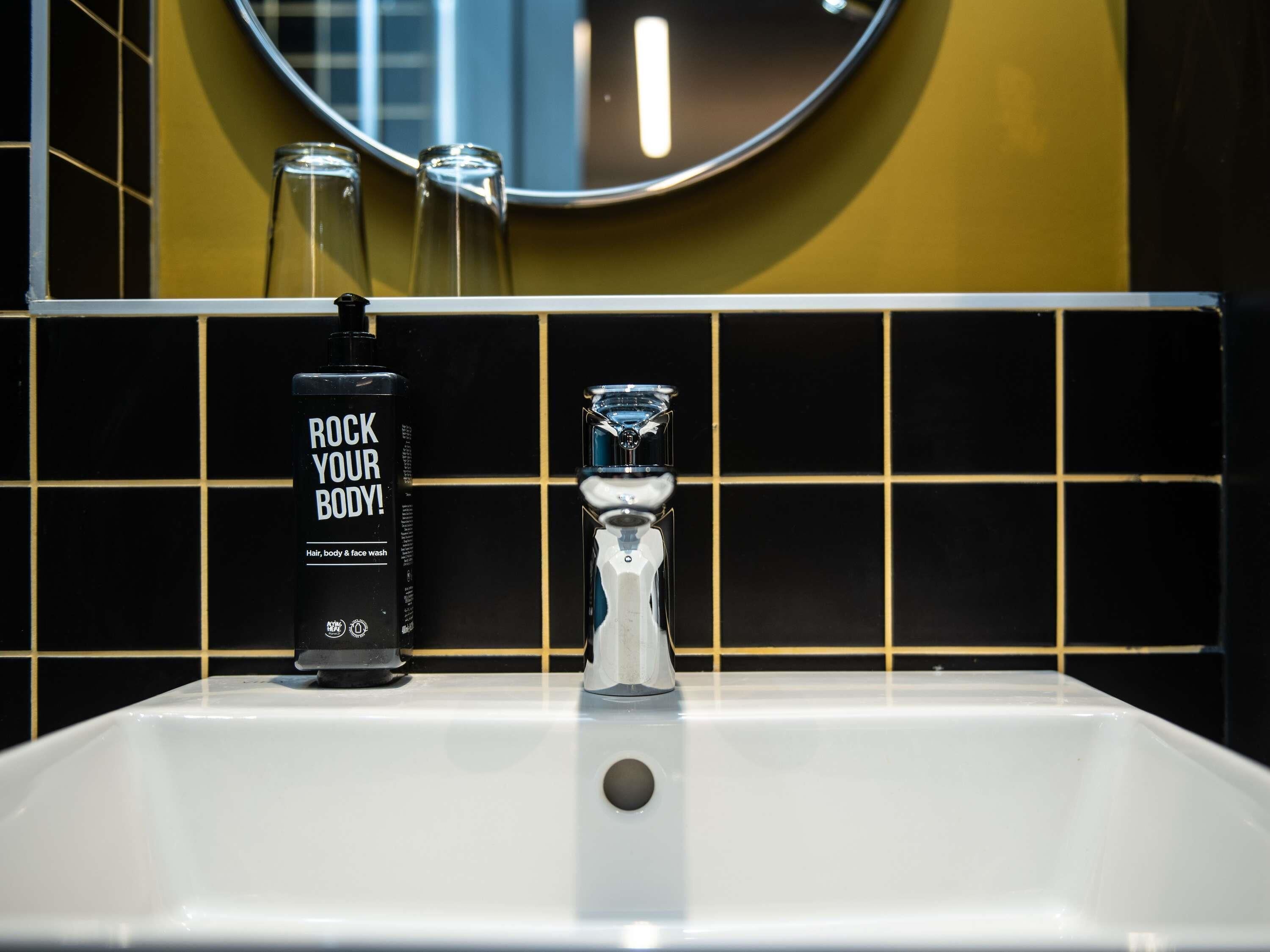 A bathroom with a white sink, a black bottle labeled 'ROCK YOUR BODY!', and a glass on the counter.