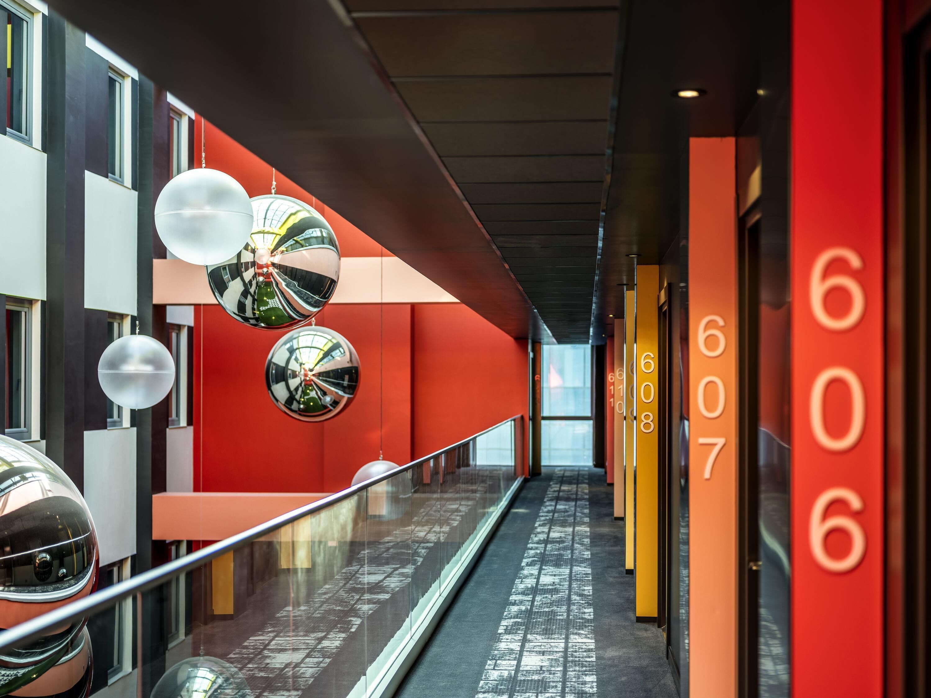 Un couloir orné de sphères décoratives réfléchissantes, doté d’une rambarde en verre et d’une série de portes portant des numéros.