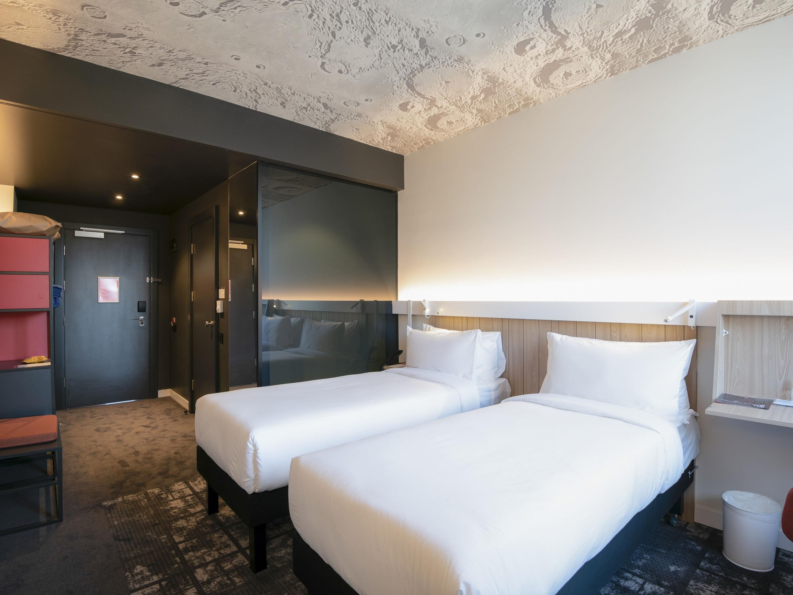 Habitación de hotel con dos camas, un escritorio, una silla, un cesto de basura y una puerta.
