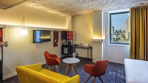 Una habitación de hotel con diseño moderno, que presenta un techo con textura distintiva, un ventanal con vistas a la ciudad y muebles contemporáneos, incluyendo un sofá amarillo y sillas rojas.