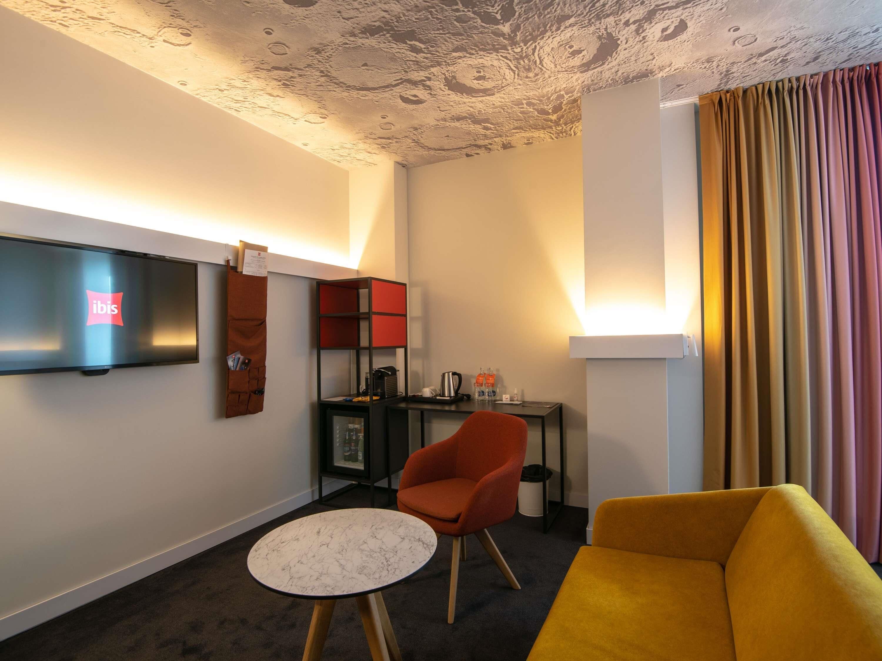 A modern hotel room with a flat-screen TV, a red chair, a yellow sofa, a small round table, and a unique ceiling design.