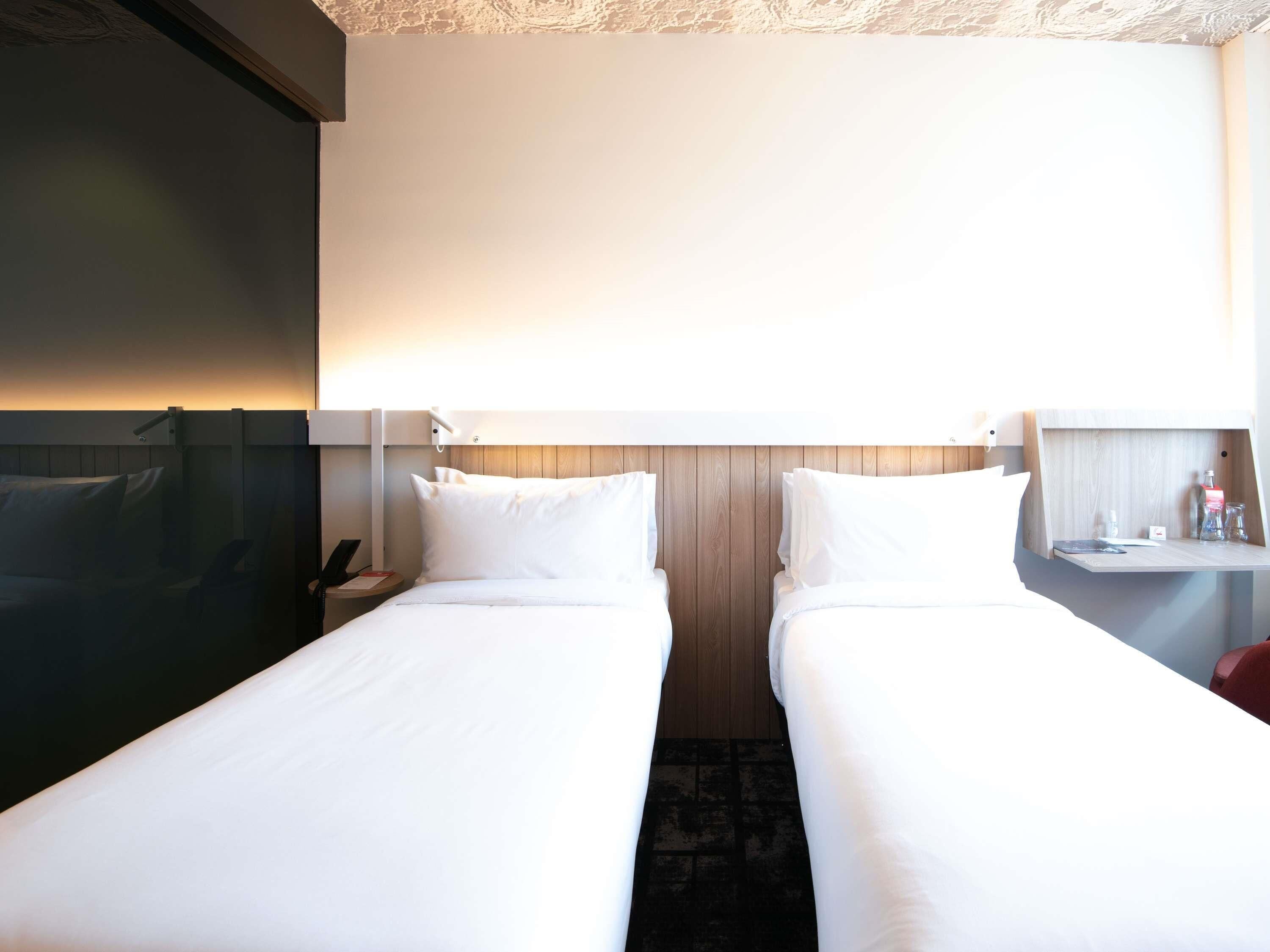 A hotel room with two beds, a desk, a nightstand, and a wall-mounted TV.