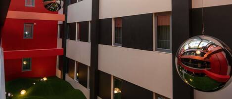 Un edificio de varios pisos con fachada roja y blanca, múltiples ventanas y un área de césped artificial verde con luces decorativas.