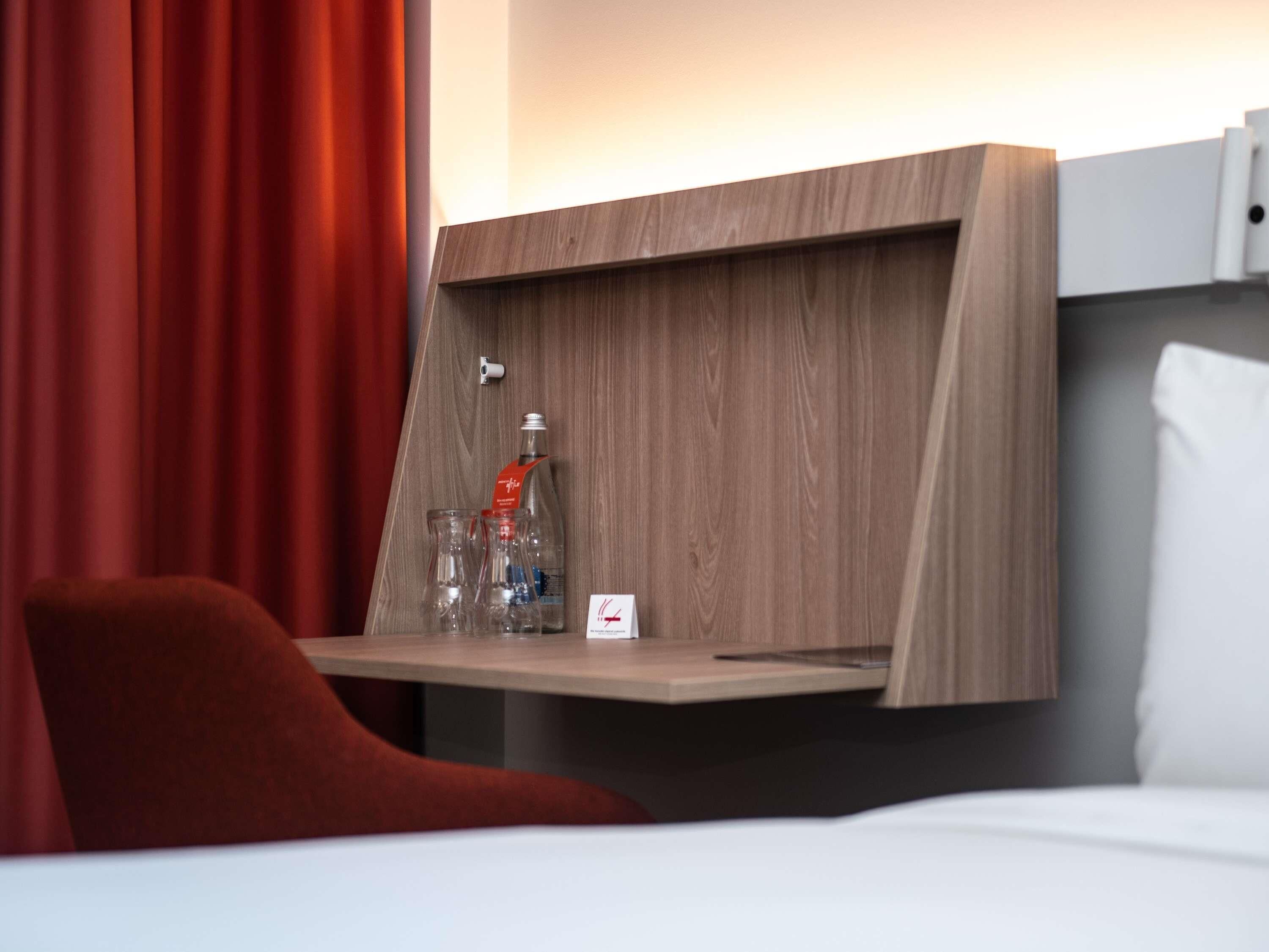 A hotel room with a wooden headboard, a bedside table with a bottle and glasses, a red chair, and a white pillow.