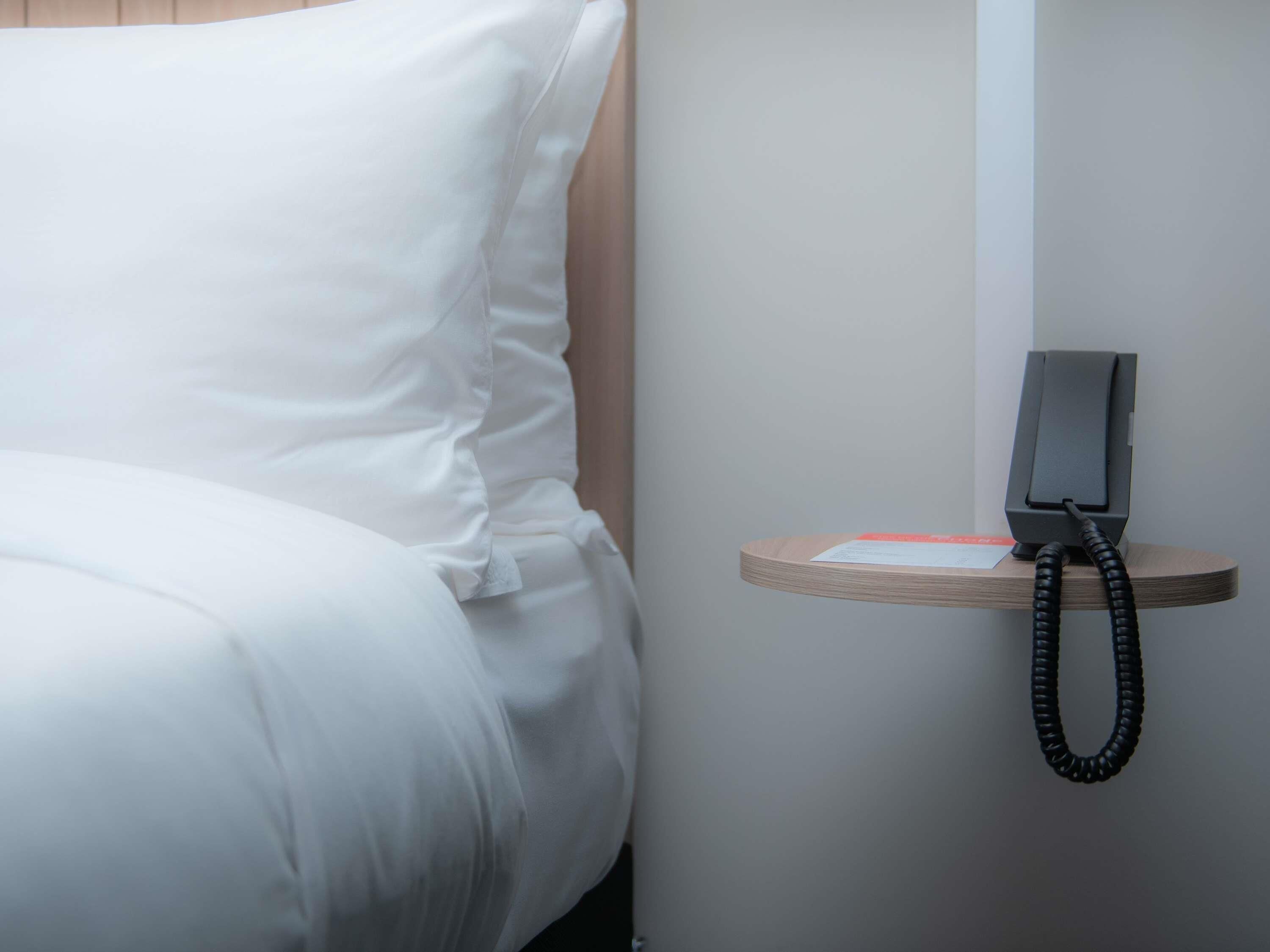 A neatly made bed with white linens, a bedside table with a telephone, and a small card or note on the table.