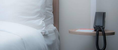 A neatly made bed with white linens, a bedside table with a telephone, and a small card or note on the table.
